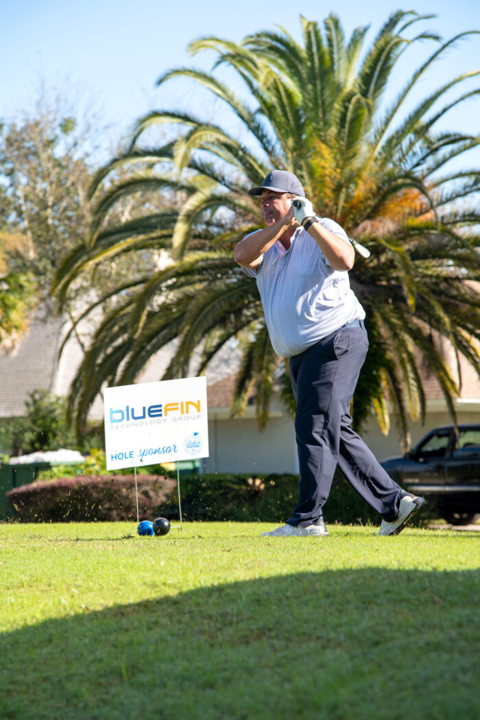 A male golfer teeing off under a sunny sky with a Bluefin Technology Group sponsor sign in the background.