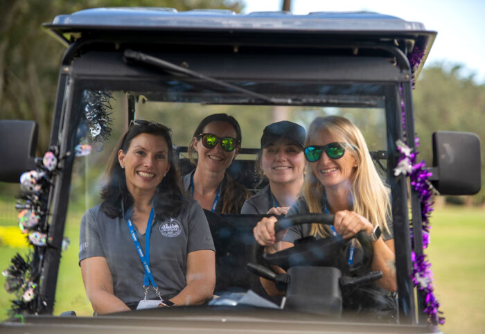 Four smiling volunteers sitting in a golf cart decorated with festive garland during the Money Pages Charity Golf Classic.
