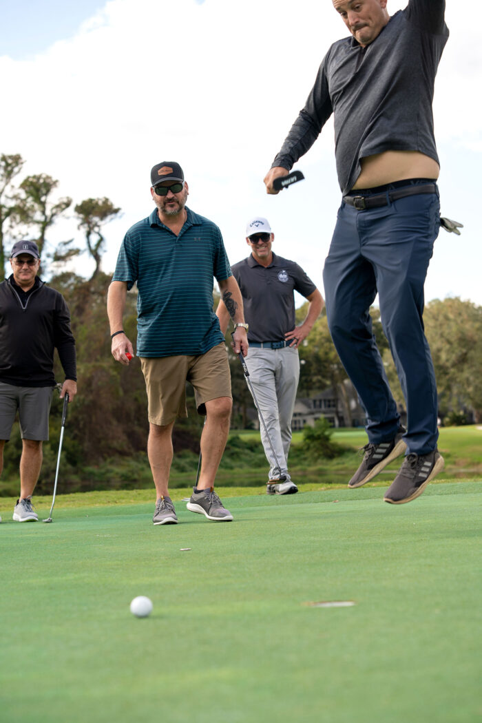 Golfer enthusiastically jumping on the green as other participants watch during the Money Pages Charity Golf Classic event.