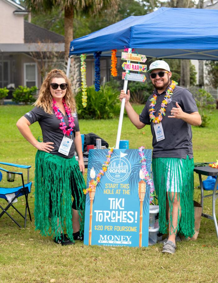 Two volunteers dressed in Hawaiian-themed attire at the tiki torch booth for the Money Pages Charity Golf Classic fundraiser.