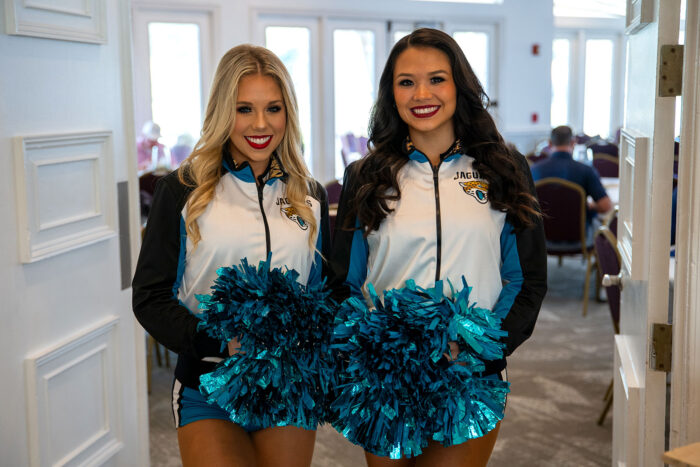 Two cheerleaders from the Jacksonville Jaguars smiling and posing indoors with teal pom-poms at the Money Pages Charity Golf Classic.